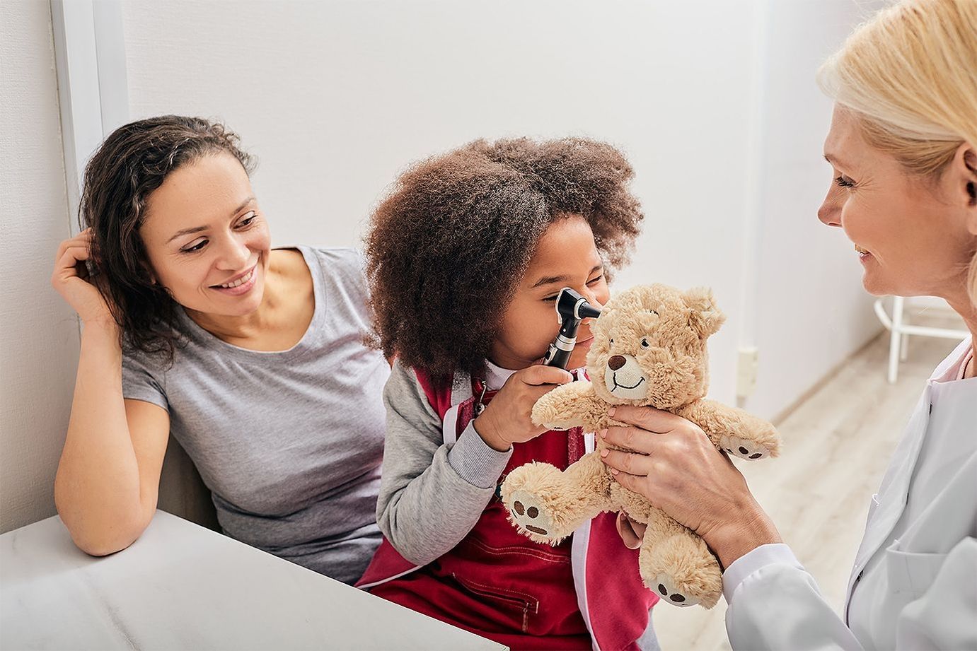 A child examines a teddy bear with an otoscope while a doctor smiles and the child's mother also smiles.